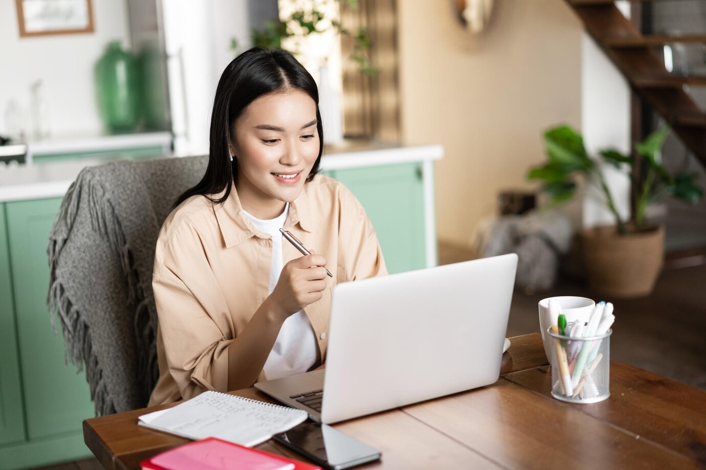 Smiling asian girl studying at home on laptop listening webinar and taking notes writing down homewo...
