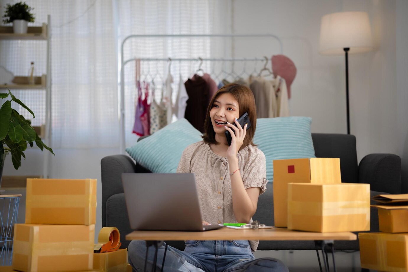 Smiling happy young Asian woman entrepreneur receiving phone call for new sales order among boxes